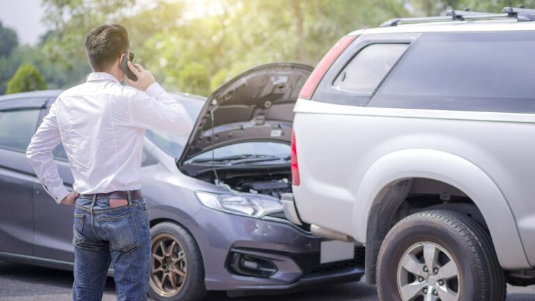 A man speaks on his cell phone while standing by an open car hood, indicating a situation after a car accident in the UAE